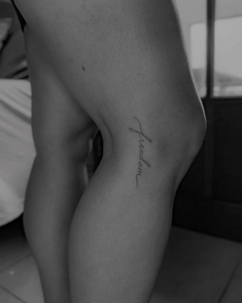 Handwritten word "freedom" lettering tattoo located on the knee.