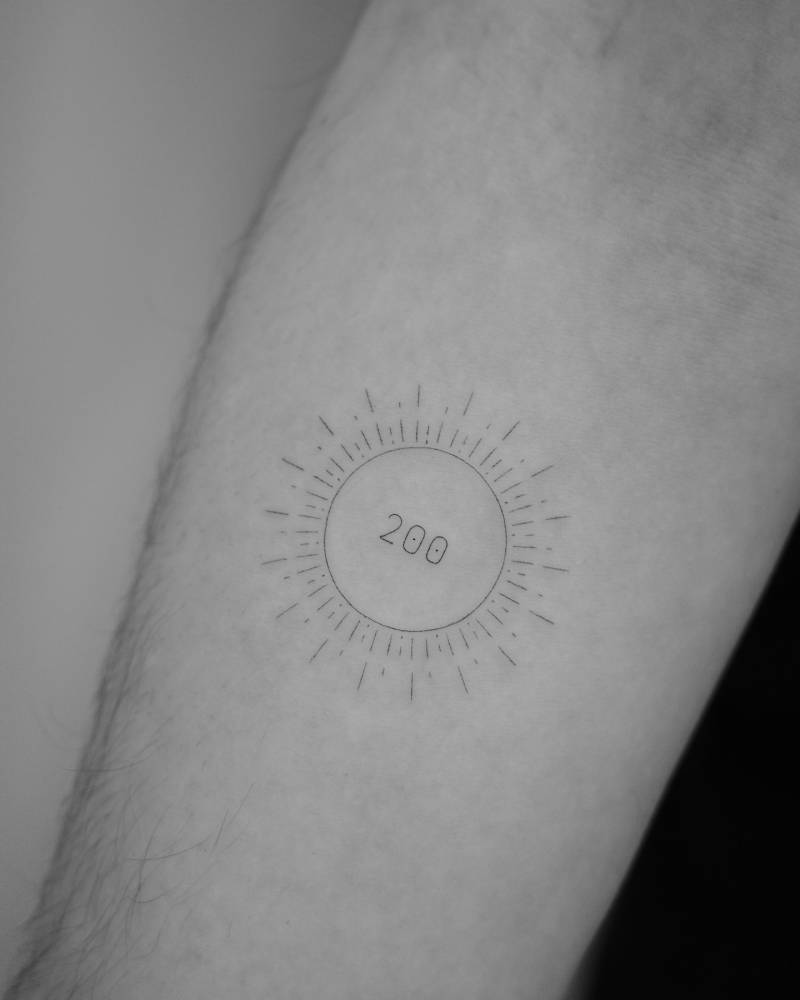 Sun and number "200" tattoo in fine line style located on the inner forearm.