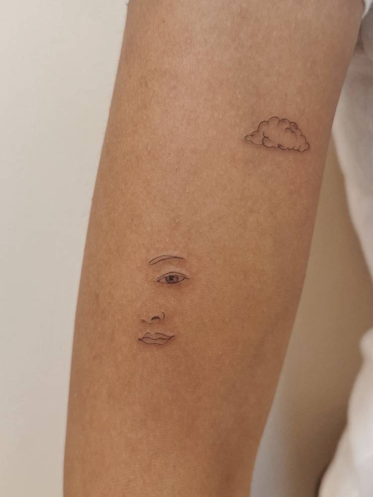 Fine line style face and cloud tattoos located on the upper arm.