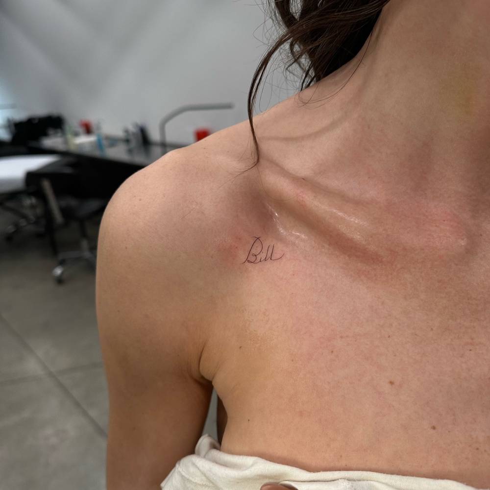 Handwritten lettering tattoo of the name "Bill" located on the collarbone.