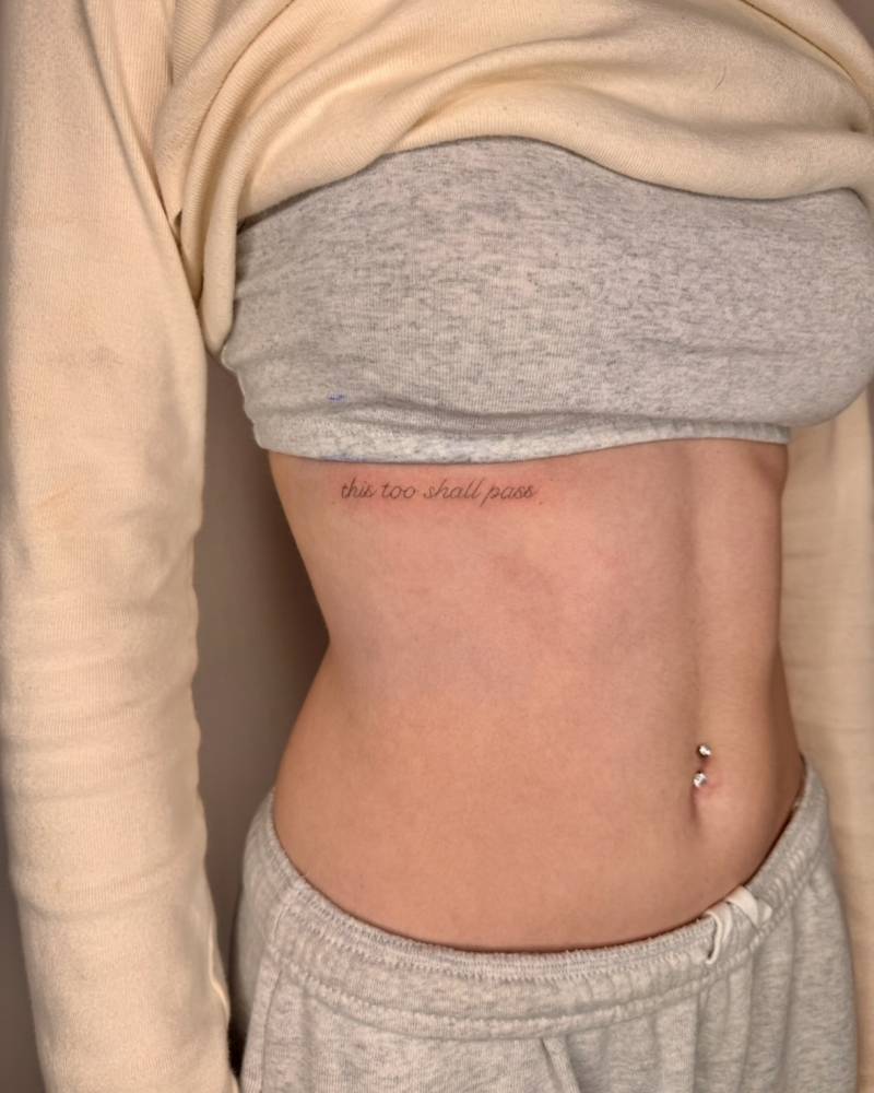 Handwritten font "this too shall pass" lettering tattoo placed on the rib.