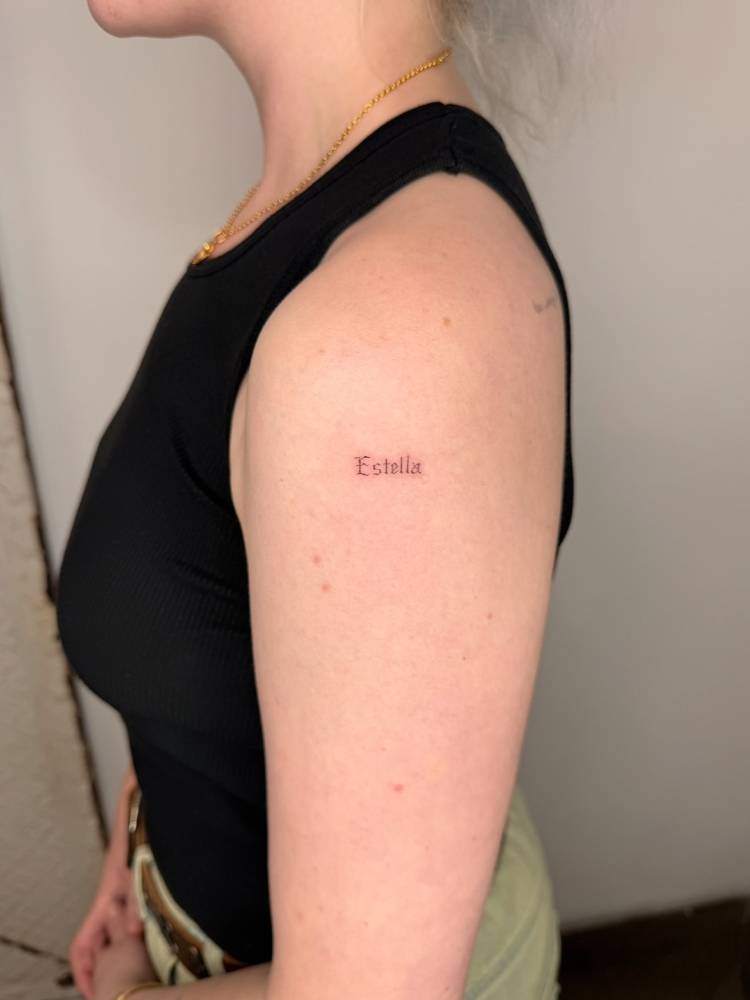"Estella" lettering tattoo in gothic font located on the upper arm.