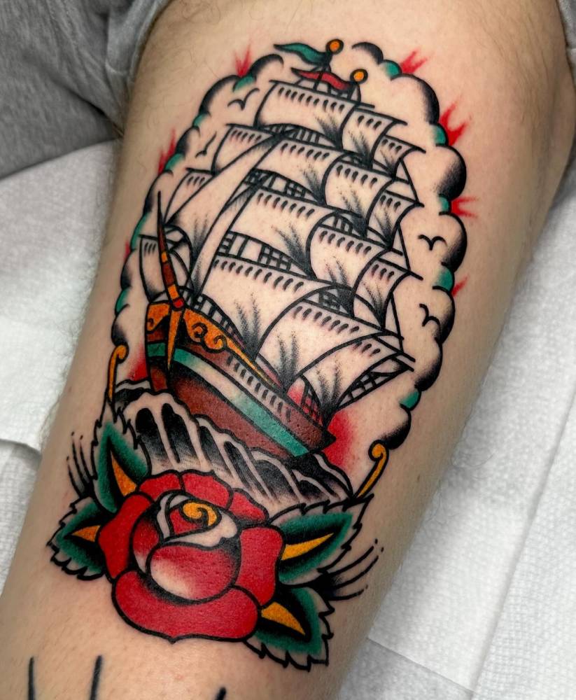 Sailing ship and red rose tattoo located on the thigh, done in traditional style.