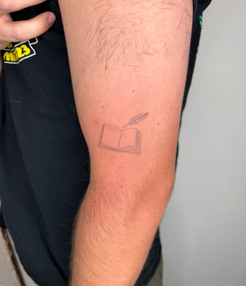 Little book and feather tattoo placed on the upper arm, done in fine line style.