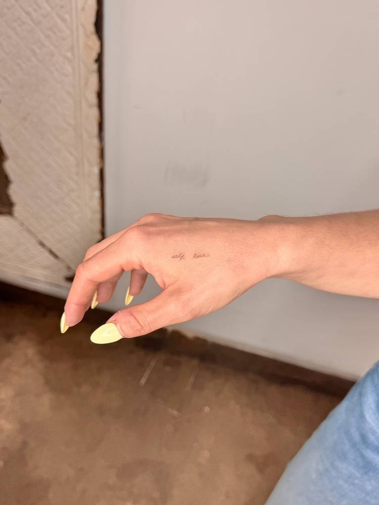"Self love" lettering tattoo located on the hand.