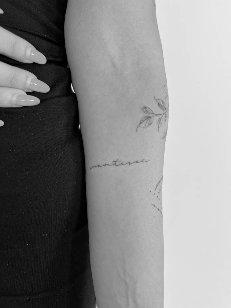Handwritten lettering tattoo of the word "ventisei" located on the inner forearm.