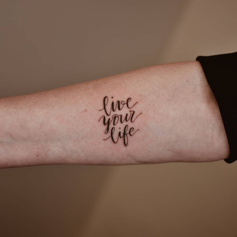 Script font "live your life" lettering tattoo done on the inner forearm.