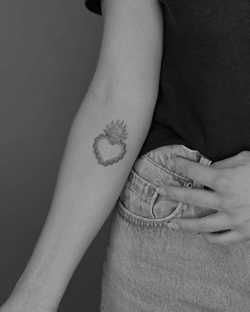 Flaming heart tattoo in illustrative style located on the inner forearm.