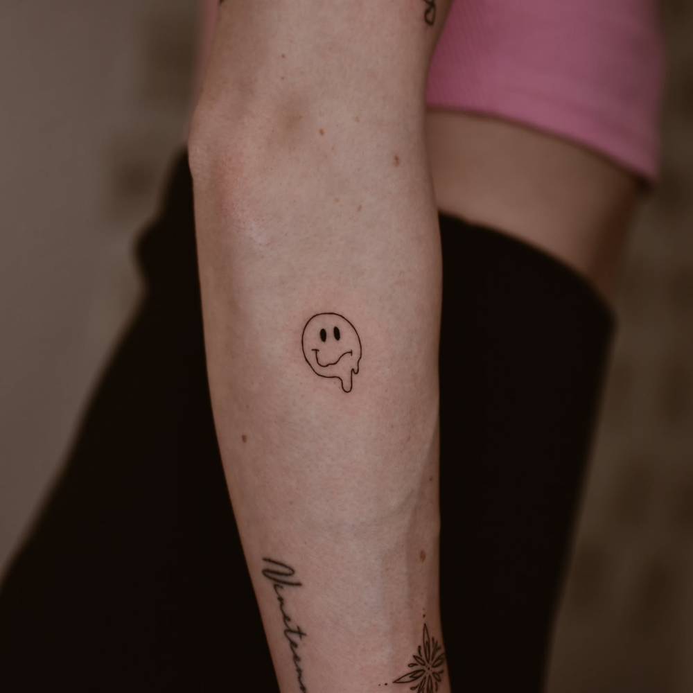 Melting smiley face tattoo in minimalistic style placed on the forearm.