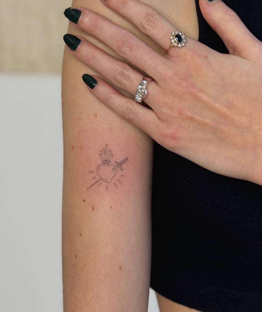 Flaming heart and sword tattoo in fine line style placed on the upper arm.