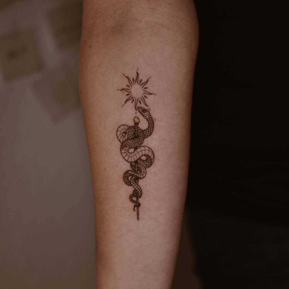 Sun, snake and sword tattoo located on the inner forearm, done in fine line style.