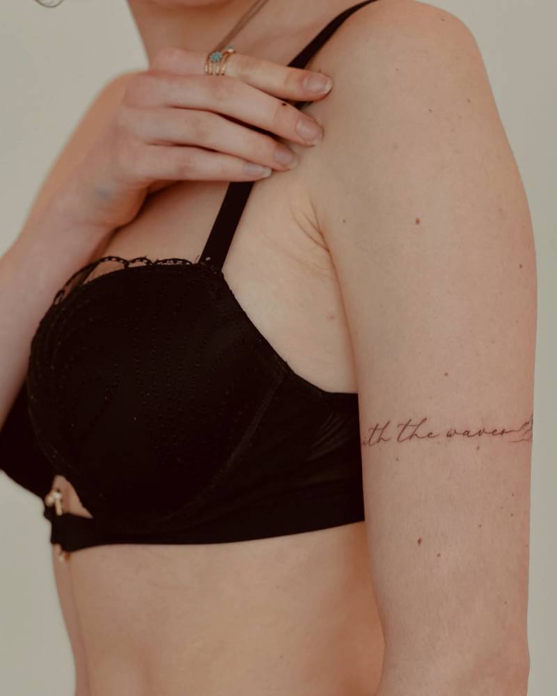 Handwritten lettering tattoo that says "With the waves" located on the upper arm.