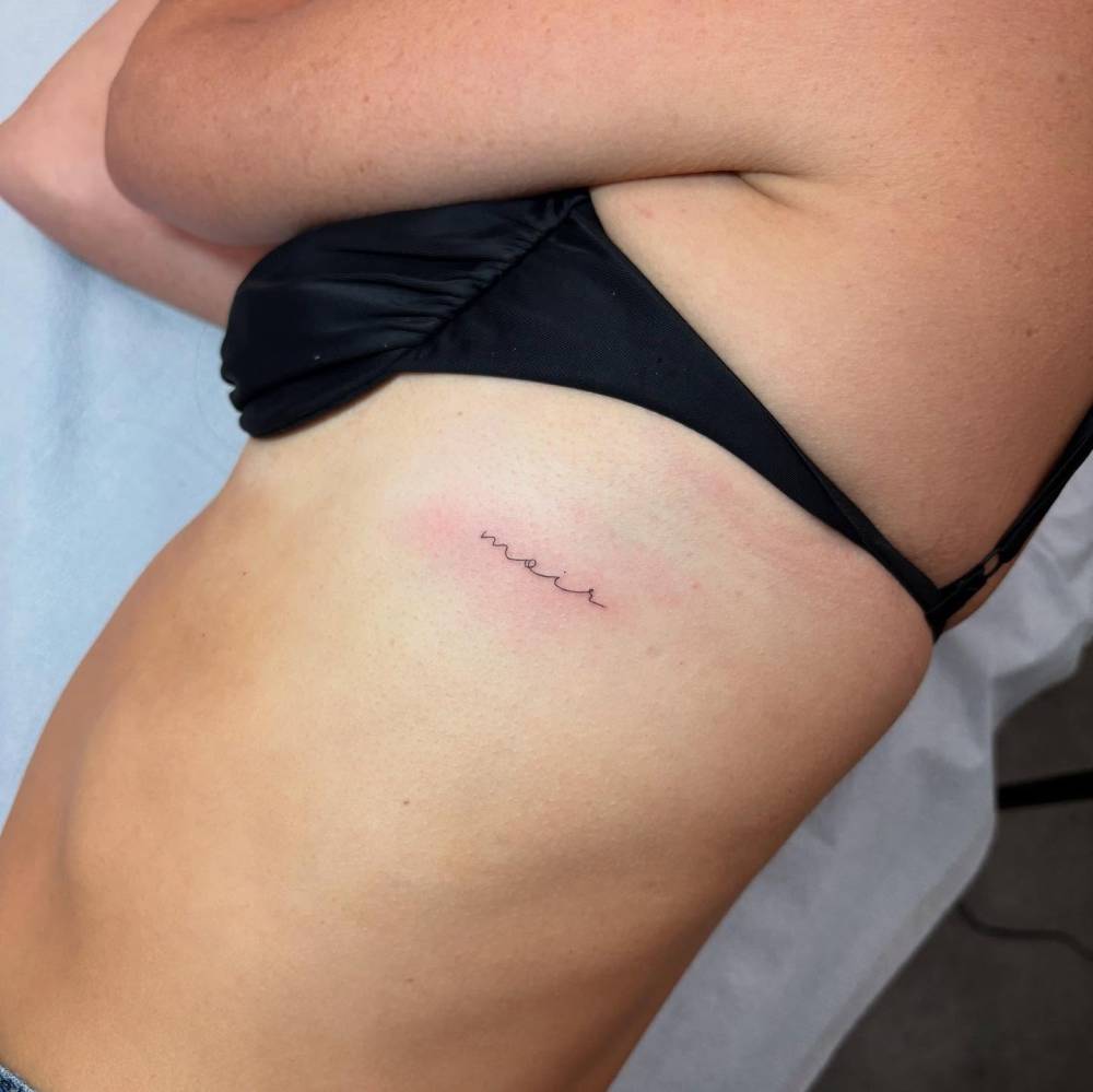 Handwritten lettering tattoo of the name "Moir" located on the rib.