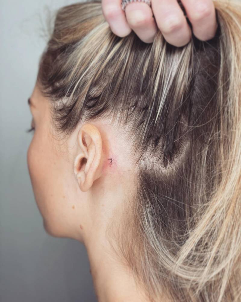 Tiny letter "G" tattoo handwritten behind the ear.