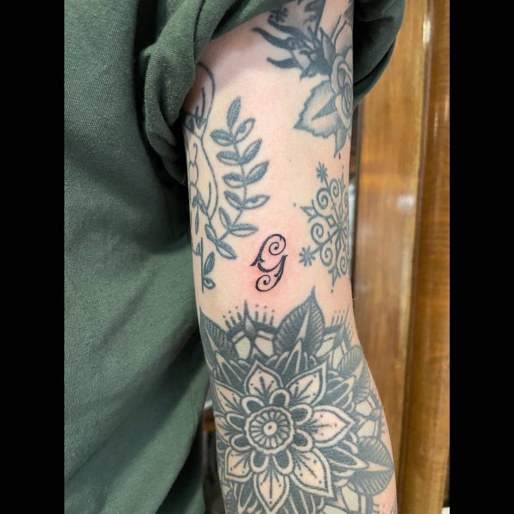 Small letter "G" gap filler tattoo located on the bicep.