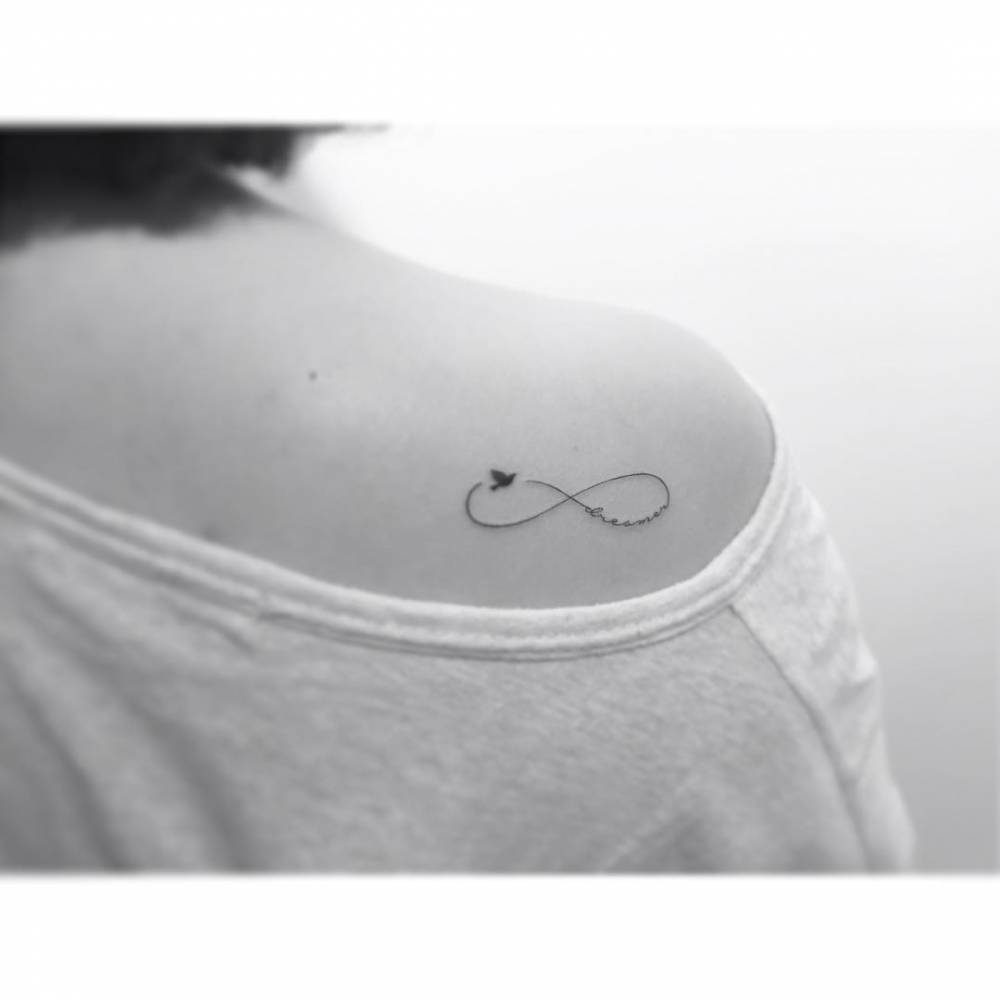 Infinity symbol, word "dreamer" and bird tattoo located on the shoulder blade.