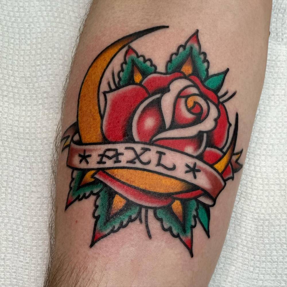 Moon, red rose and name "Axl" tattooed on the inner forearm.