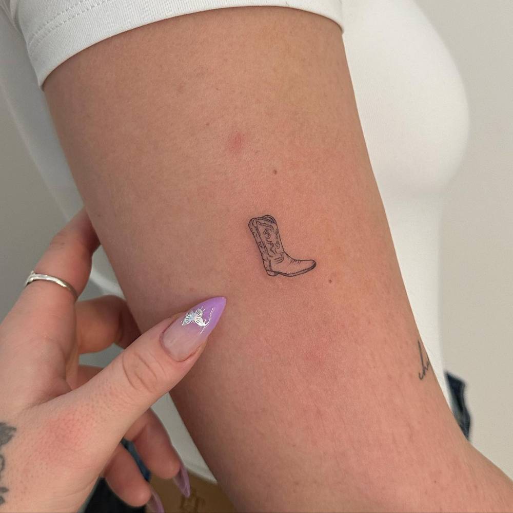 Fine line style cowboy boot tattoo located on the upper arm.