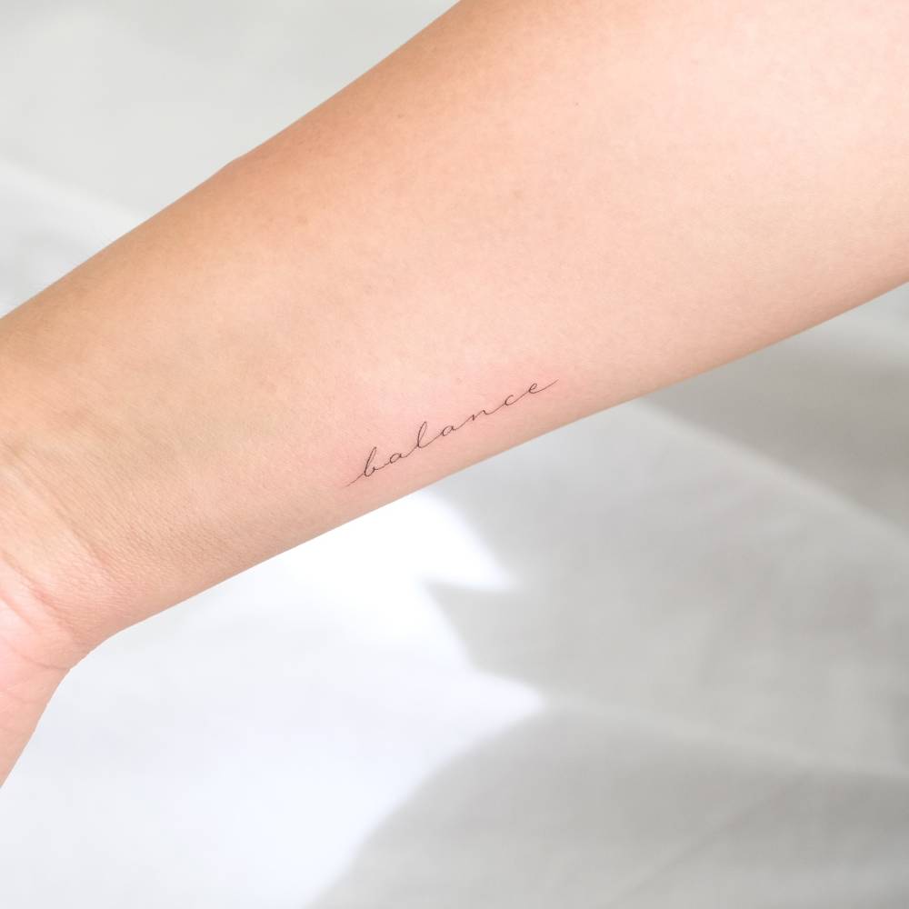Handwritten lettering tattoo of the word "balance" located on the wrist.