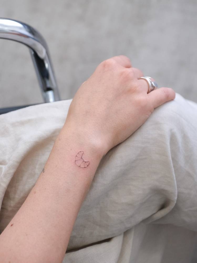 Tiny tattoo of a croissant in fine line style placed on the wrist.