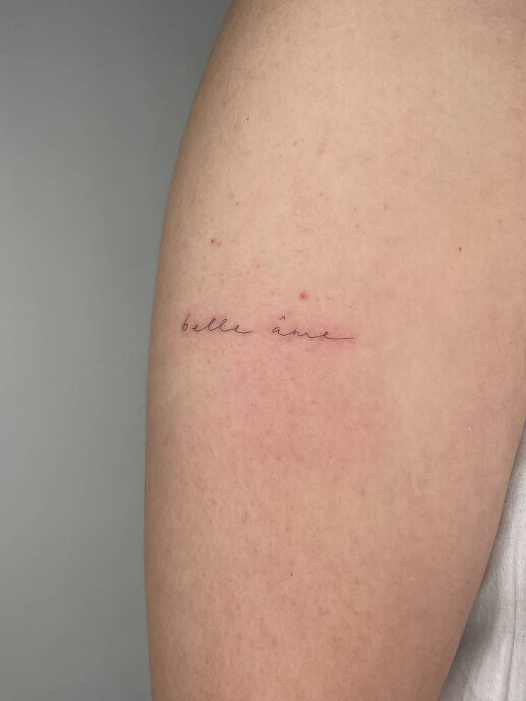 "Belle ame" lettering tattoo in handwritten font located on the upper arm.