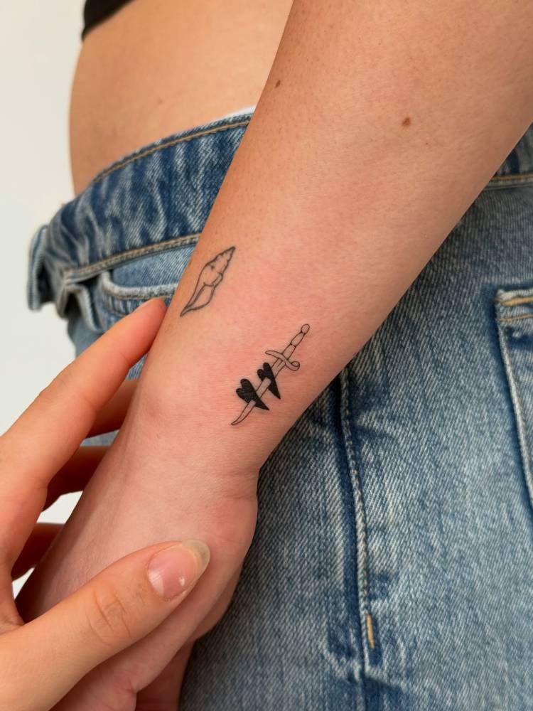 Hearts and dagger tattoo in minimalistic style located on the wrist.