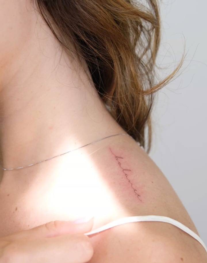 Handwritten lettering tattoo of the word "balance" placed on the top of shoulder.