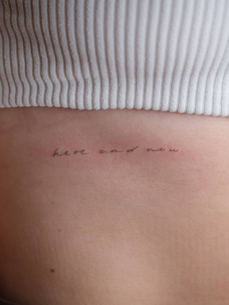 Handwritten font "here and now" lettering tattoo placed on the rib.