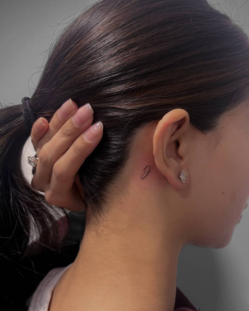 Tiny letter "J" tattoo placed behind the ear.