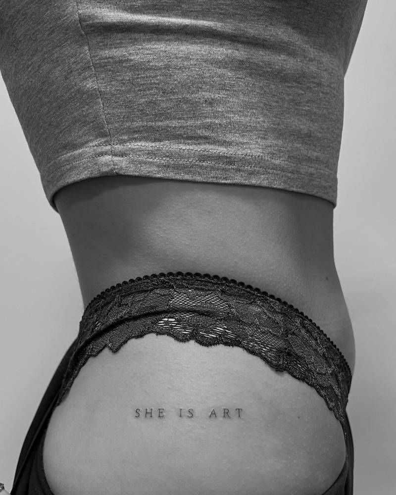 "She is art" lettering tattoo in typewriter font located on the hip.