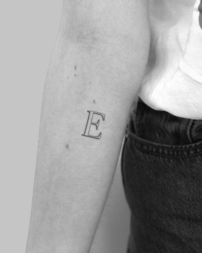 Capital letter "E" in serif font tattooed on the inner forearm.
