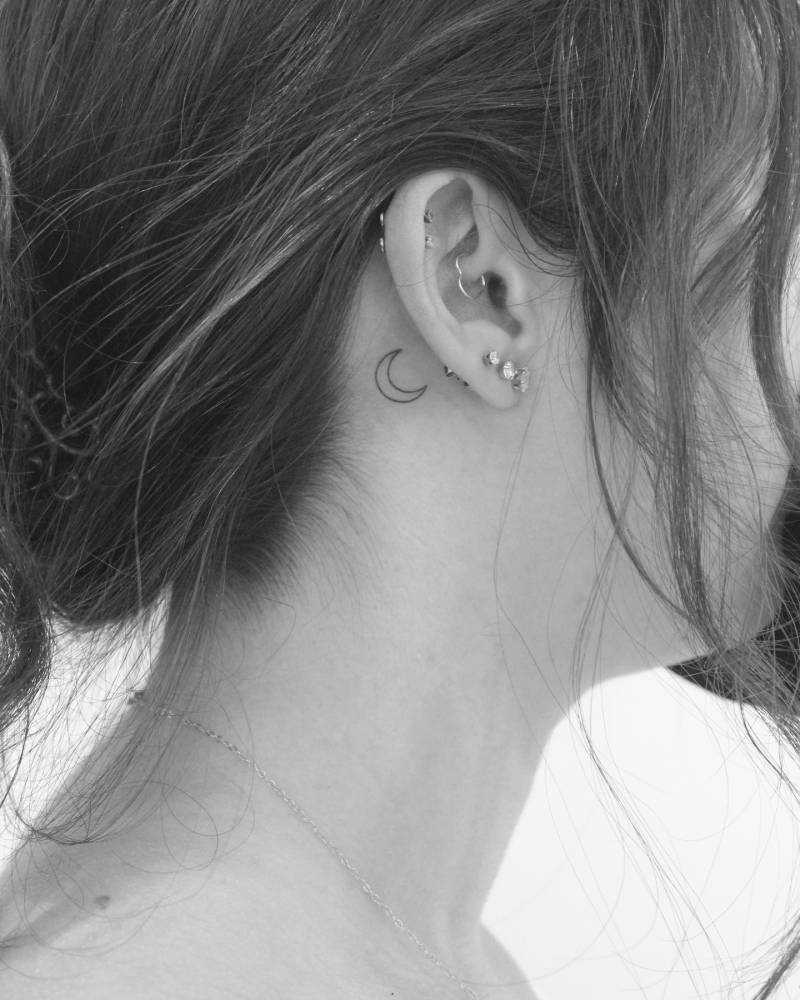 Tiny crescent moon tattoo in minimalistic style located behind the ear.