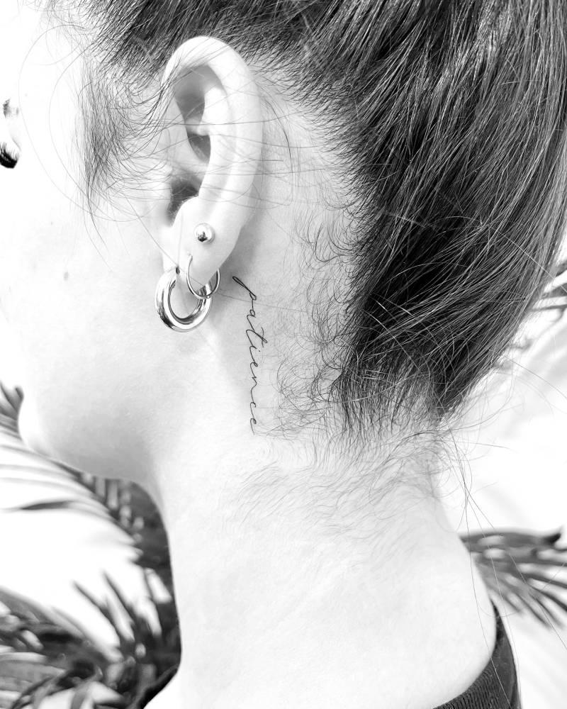 Handwritten font word "patience" tattoo located behind the ear.