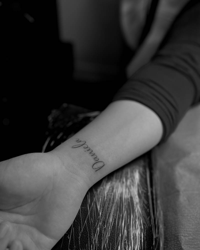 Name "Daniela" lettering tattoo located on the wrist.