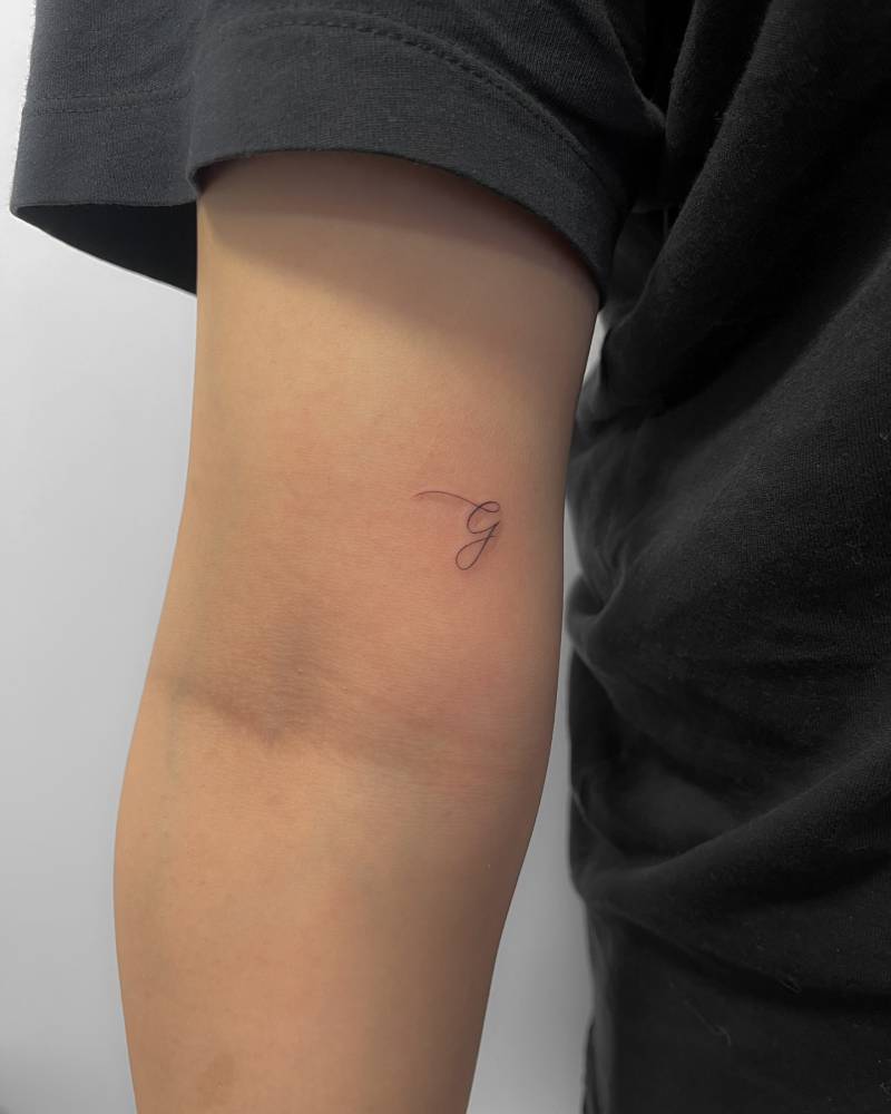 Tiny letter "G" tattoo in handwritten font located on the inner arm.