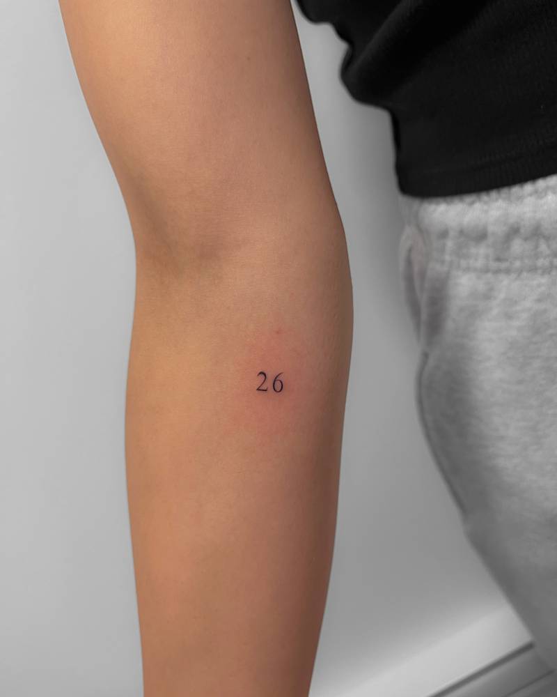 Tiny lettering tattoo of the number "26" located on the inner forearm.