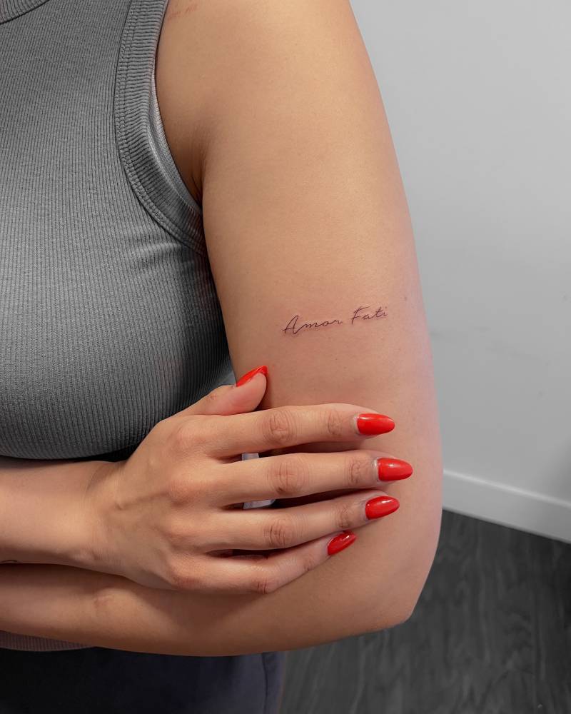 "Amor fati" lettering tattoo handwritten on the upper arm.
