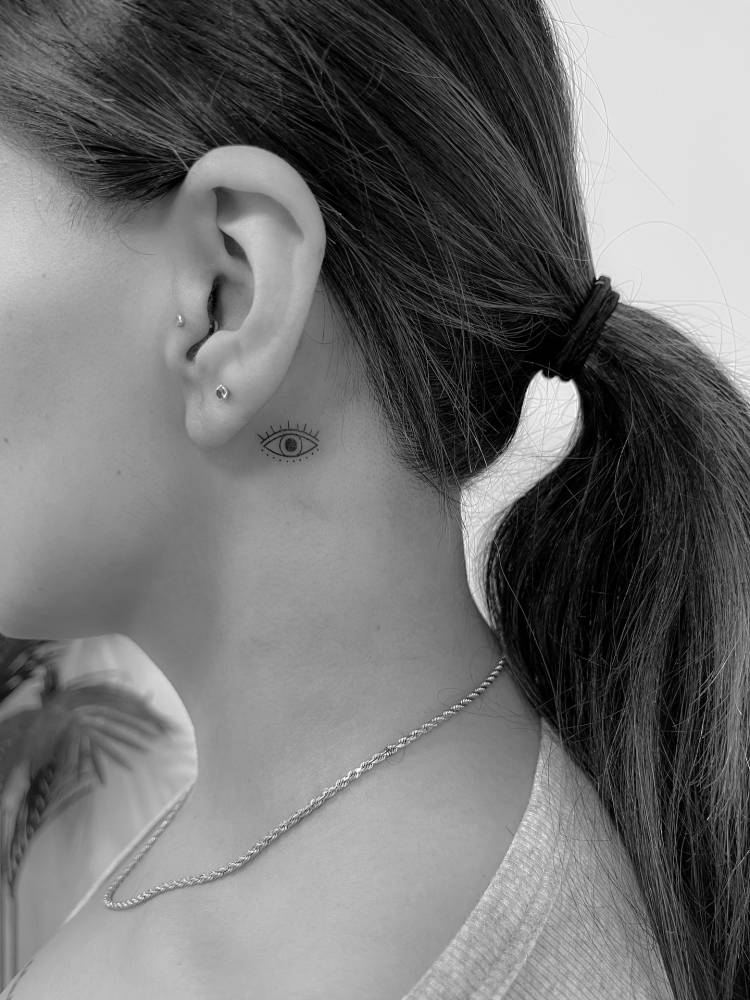 Tiny minimalistic evil eye tattoo located behind the ear.