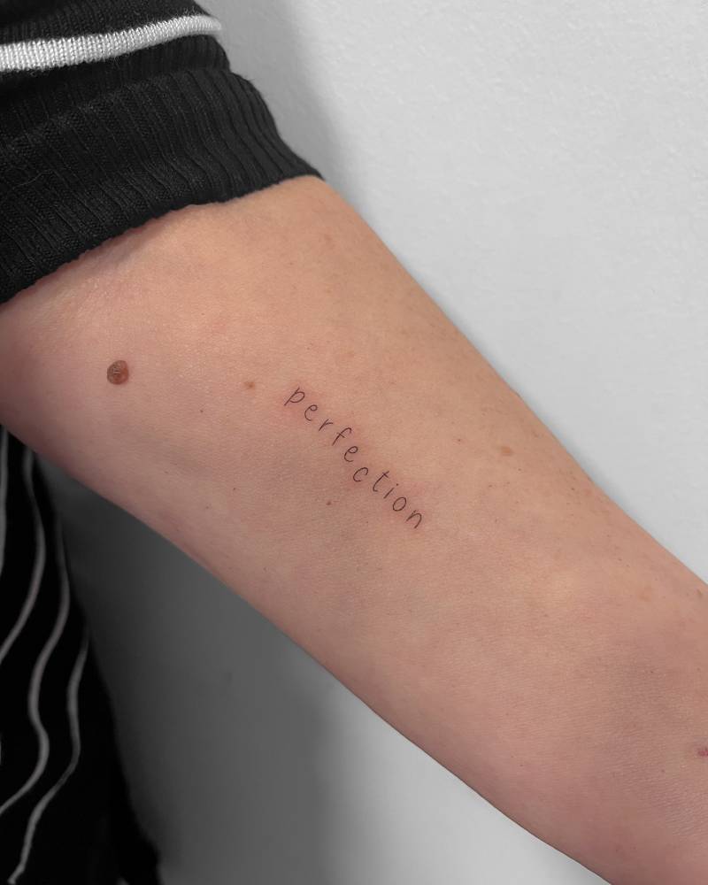 Word "perfection" lettering tattoo located on the inner forearm.
