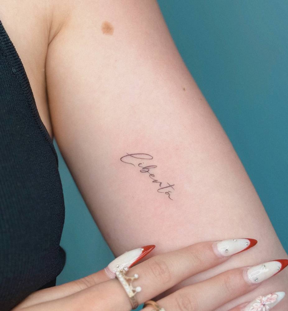 Handwritten lettering tattoo of the word "liberta" located on the inner arm.