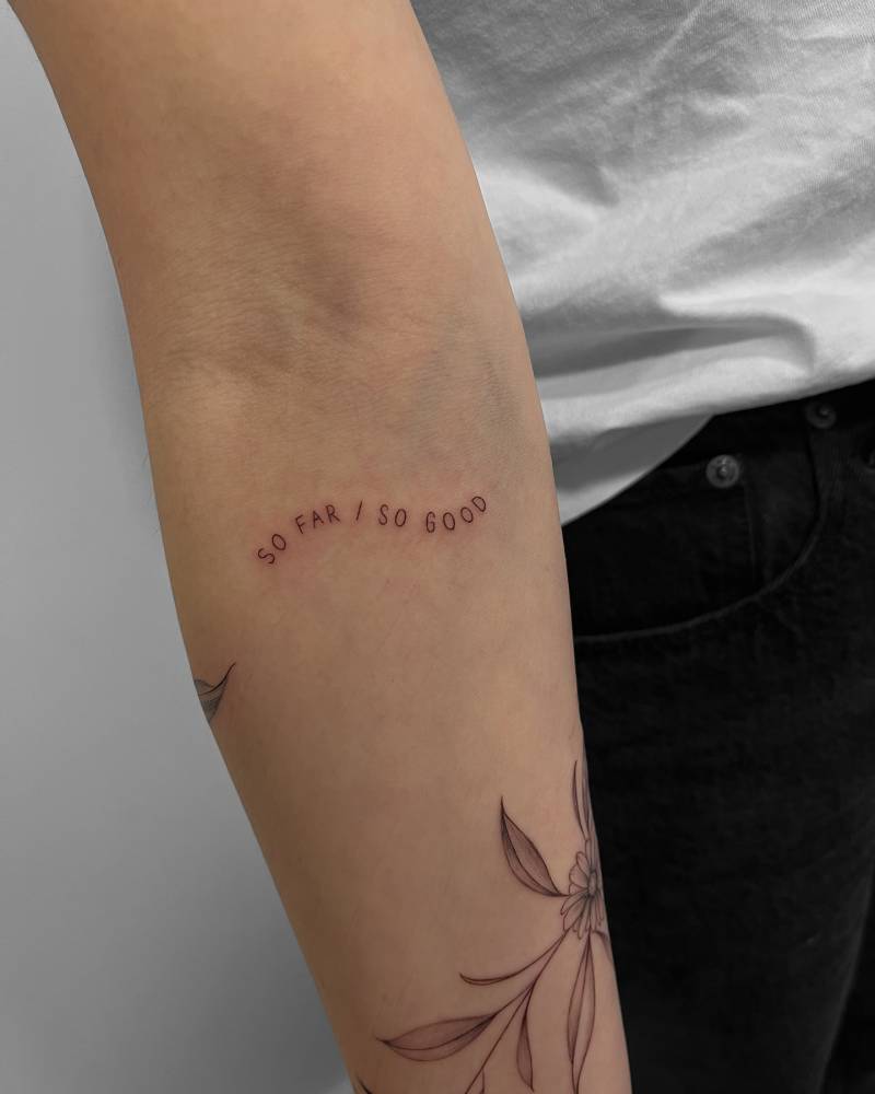 Lettering tattoo that says "so far, so good" located on the inner forearm.