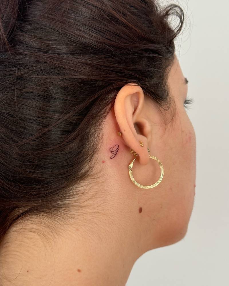 Little letter "G" tattoo placed behind the ear.