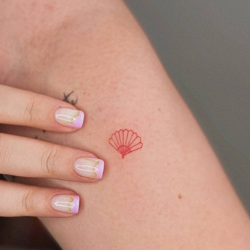 Tiny hand fan in red ink tattooed on the inner forearm, fine line style.