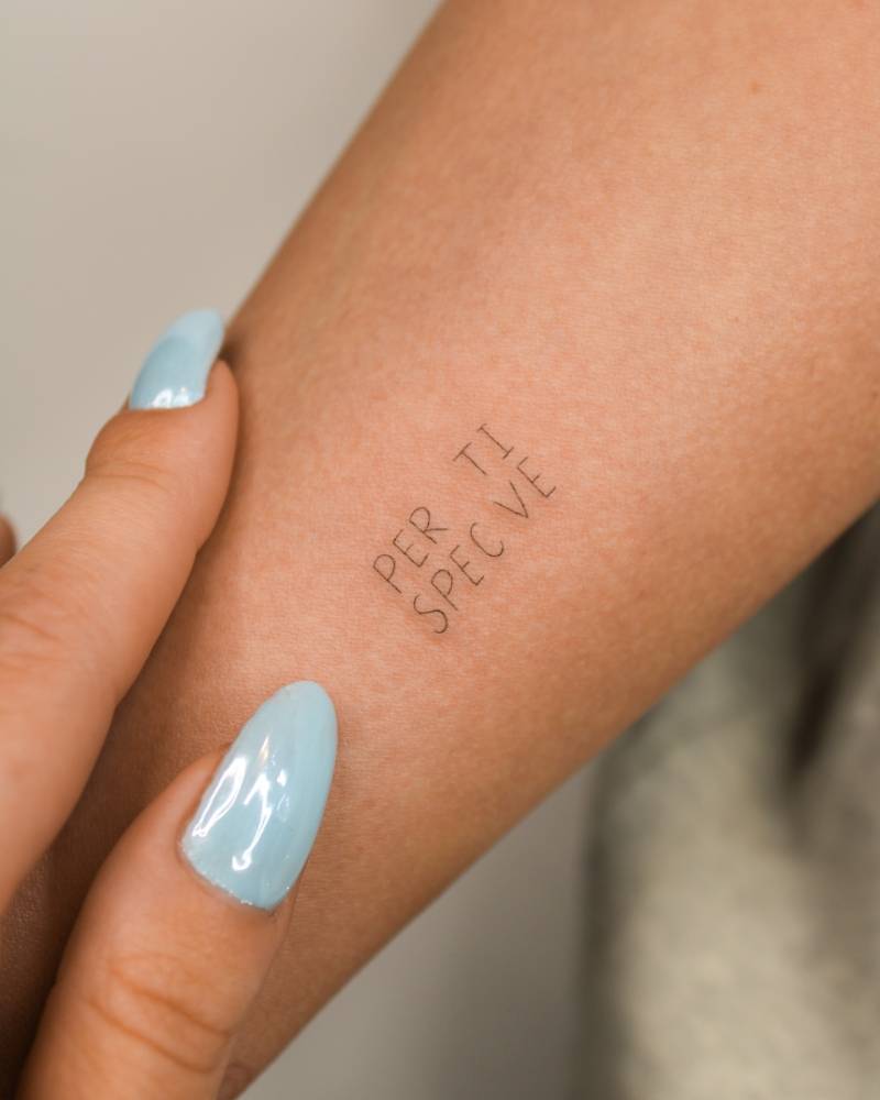 Handwritten lettering tattoo of the word "perspective" located on the inner forearm.