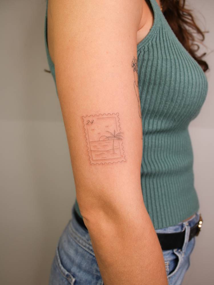 Beach stamp tattoo located on the upper arm, done in fine line style.