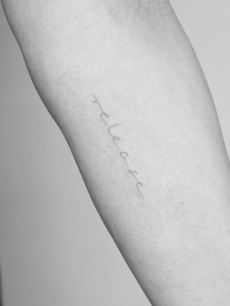 Handwritten lettering tattoo of the word "release" located on the inner forearm.