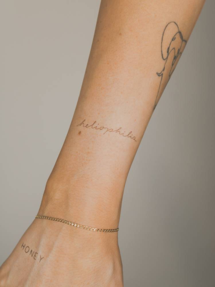 Handwritten lettering tattoo of the word "heliophilia" located on the wrist.