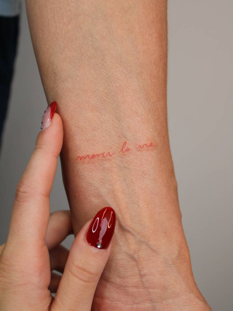 "Merci la vie" handwritten tattoo in red ink located on the wrist.