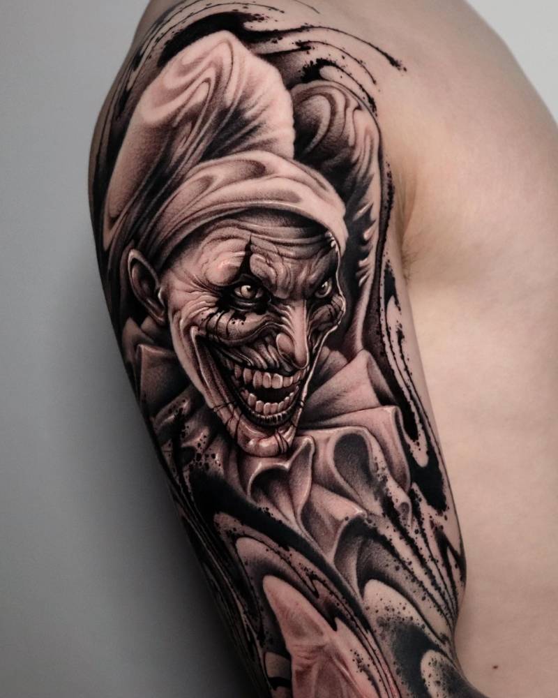 Black and grey style joker portrait tattoo located on the upper arm.