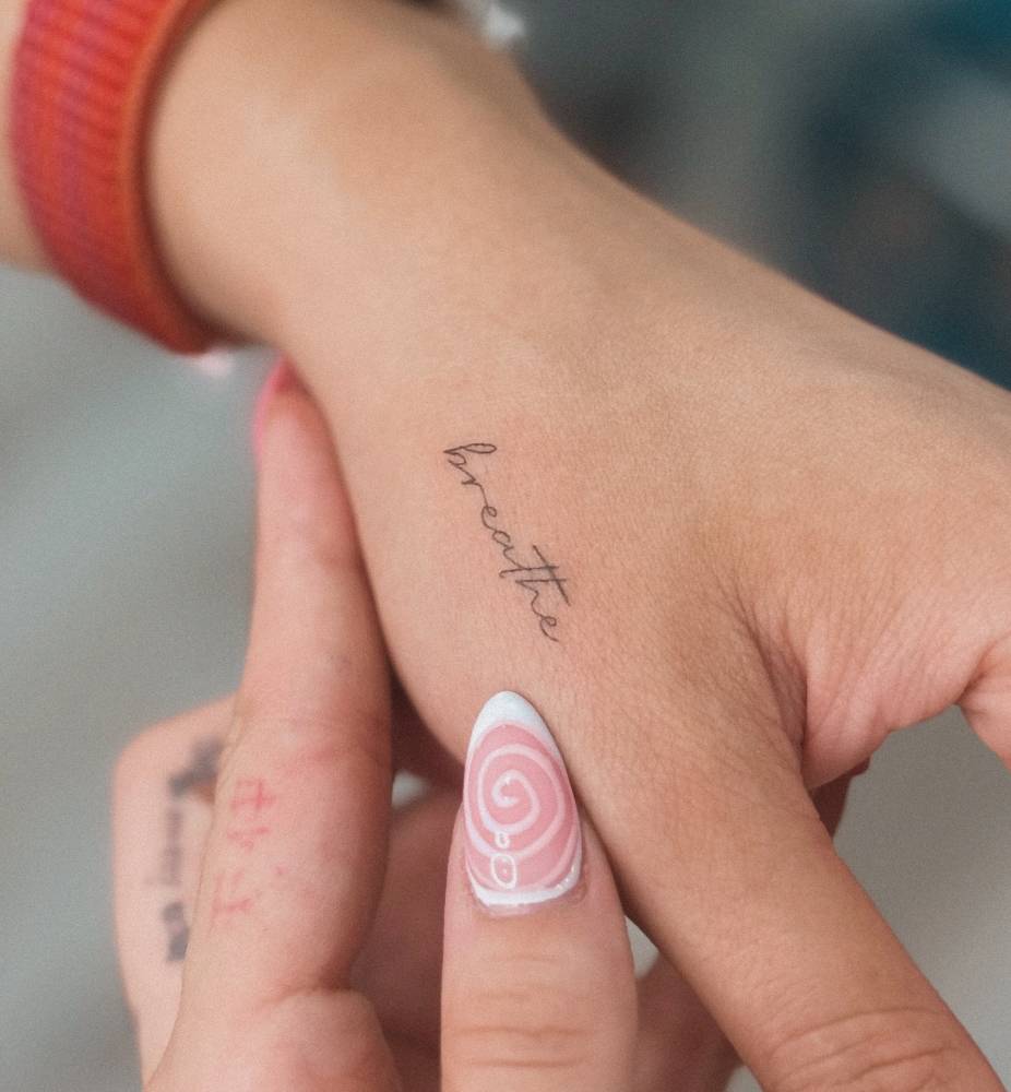 Handwritten lettering tattoo of the word "breathe" placed on the hand.